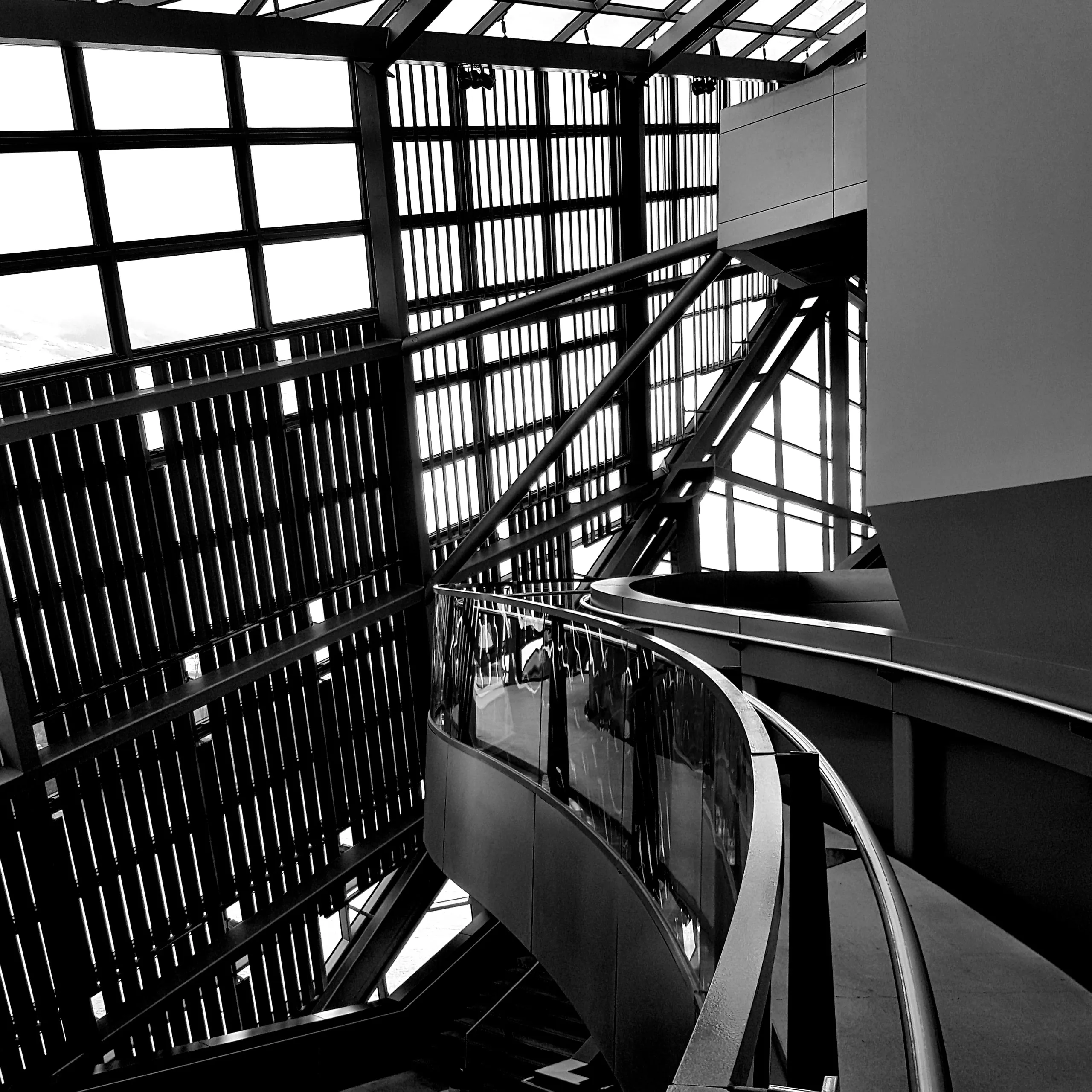 Black and white interior of a glazed hall, steel roof truss overhead and a curved balustrade sweeping through the frame