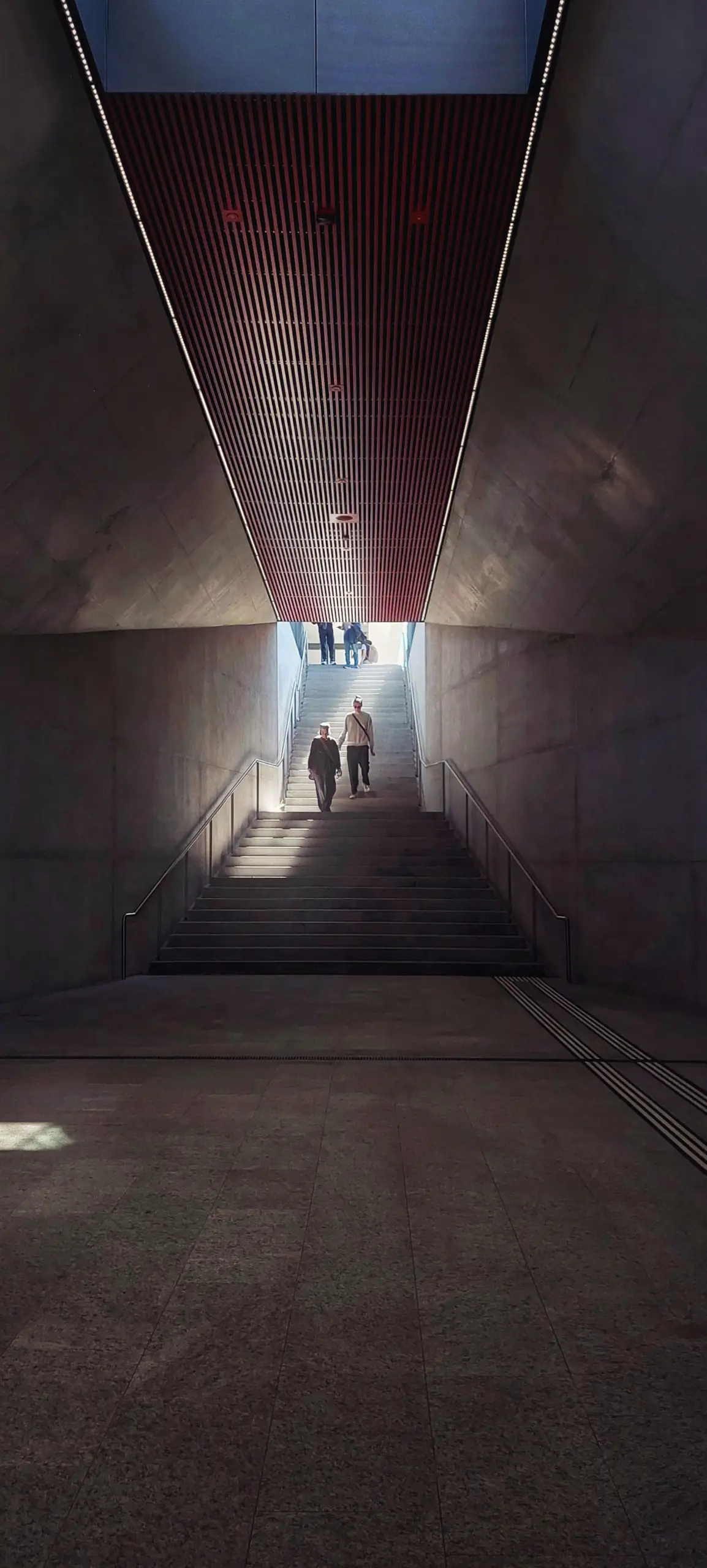 Long concrete corridor narrowing to a bright opening, two figures silhouetted at the far end