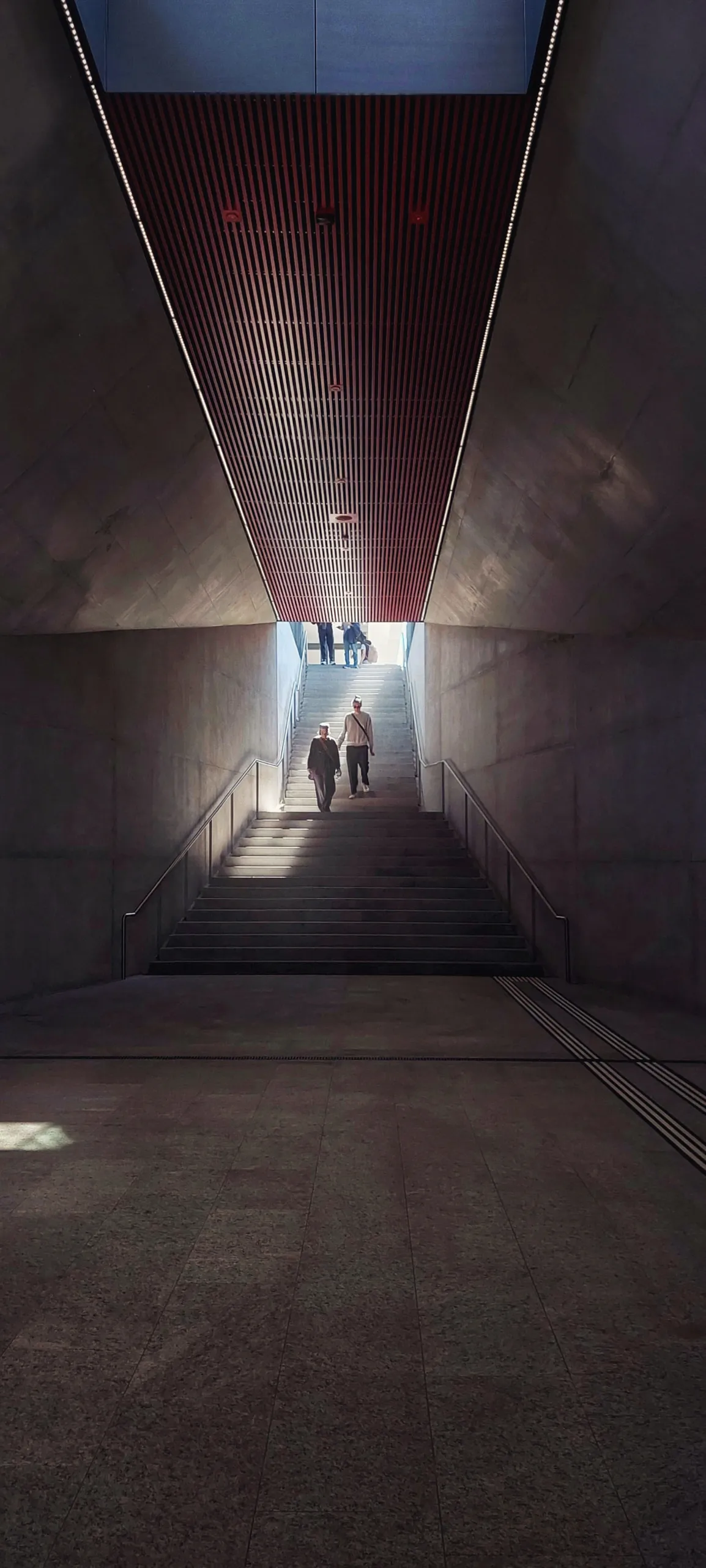 Long concrete corridor narrowing to a bright opening, two figures silhouetted at the far end