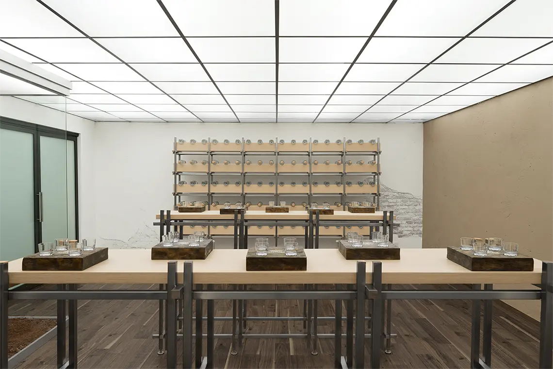 Tasting room — pale timber tables set with glassware on dark wooden trays, a wall of steel racks holding rows of glasses behind