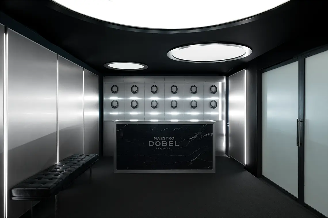 Dark reception room lined in brushed steel, a black marble desk carrying the Maestro Dobel wordmark under a grid of recessed lights