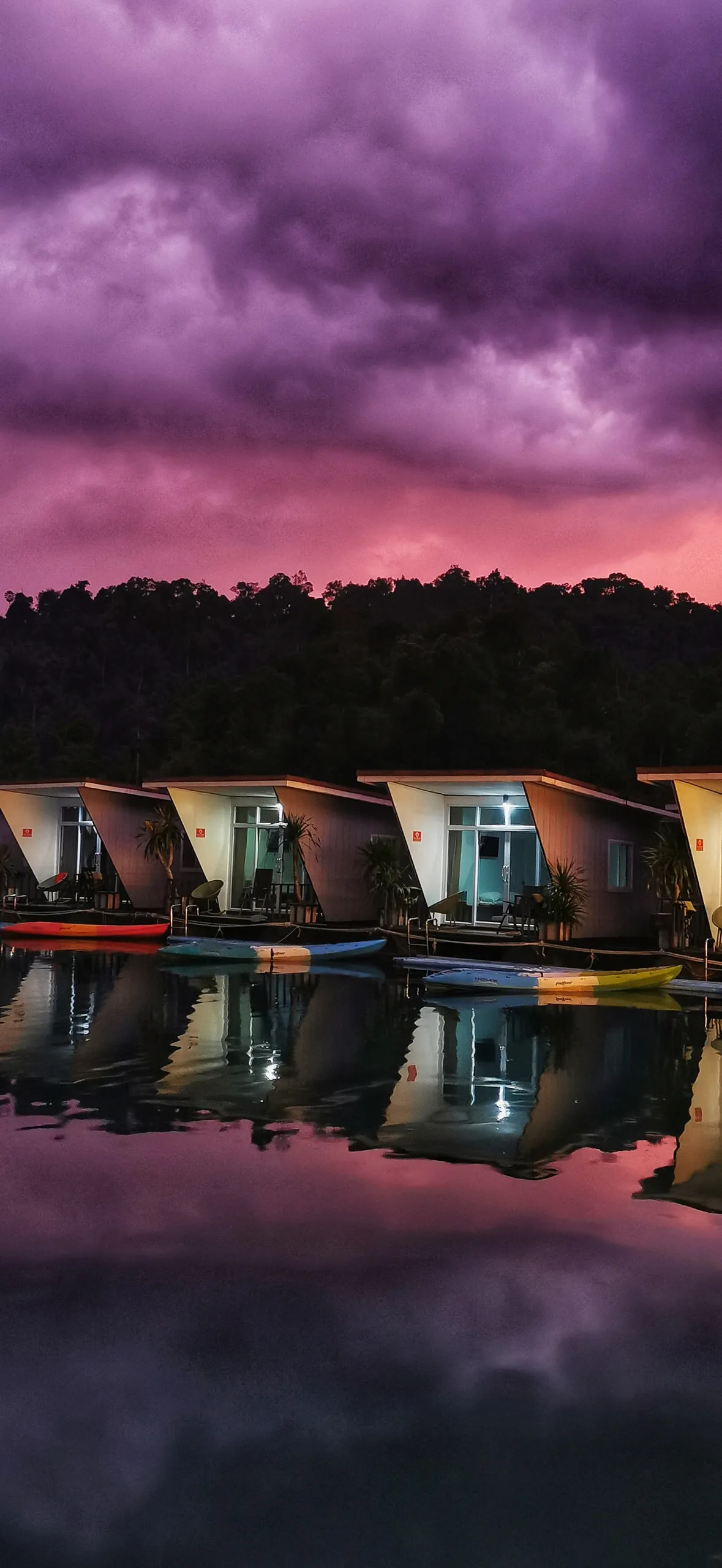 A lit modern house mirrored in still water at dusk under a violet storm sky