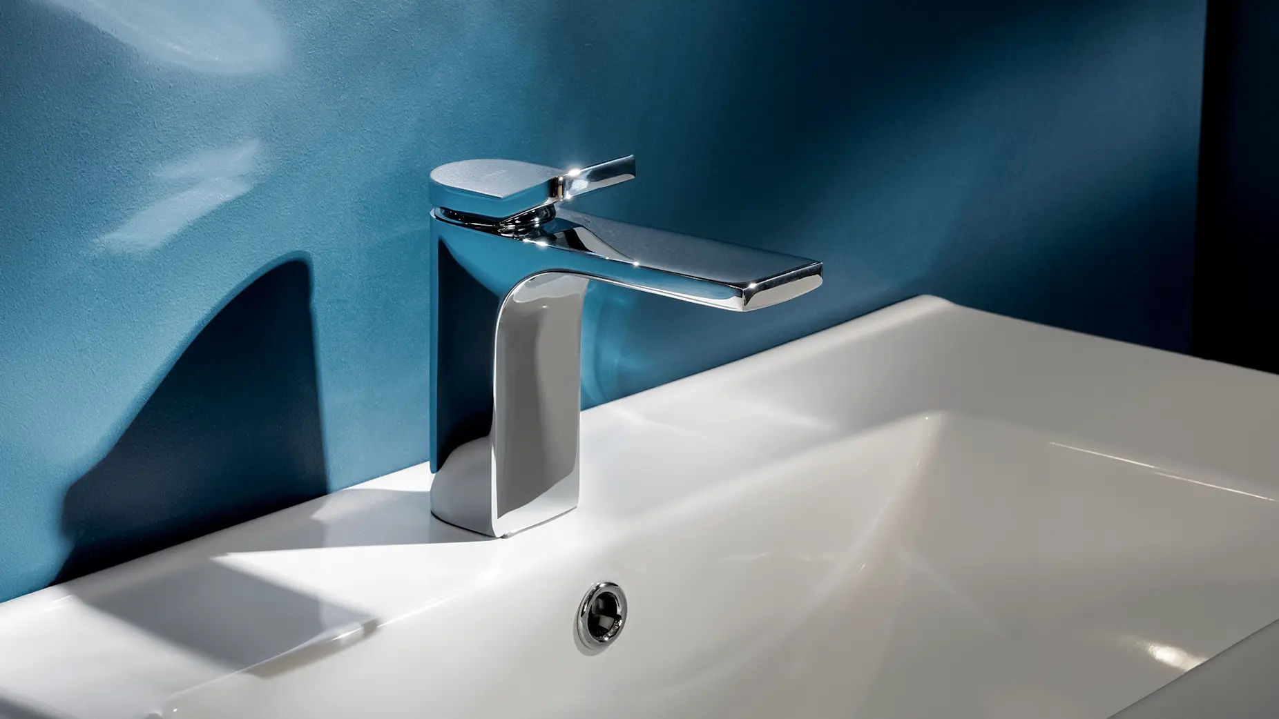 The finished mixer installed on a white ceramic basin against a deep blue plaster wall, raking light along the polished spout
