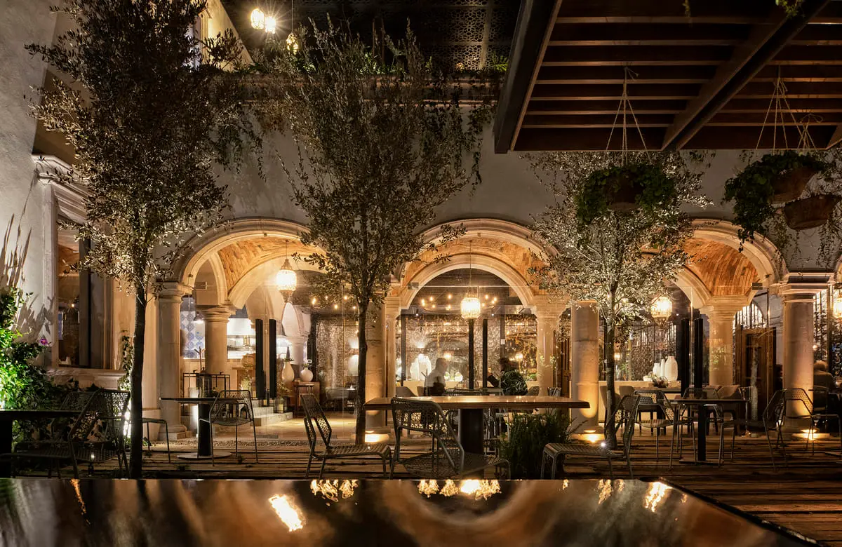 Arcaded courtyard at night, warm light under stone arches reflected in a still pool