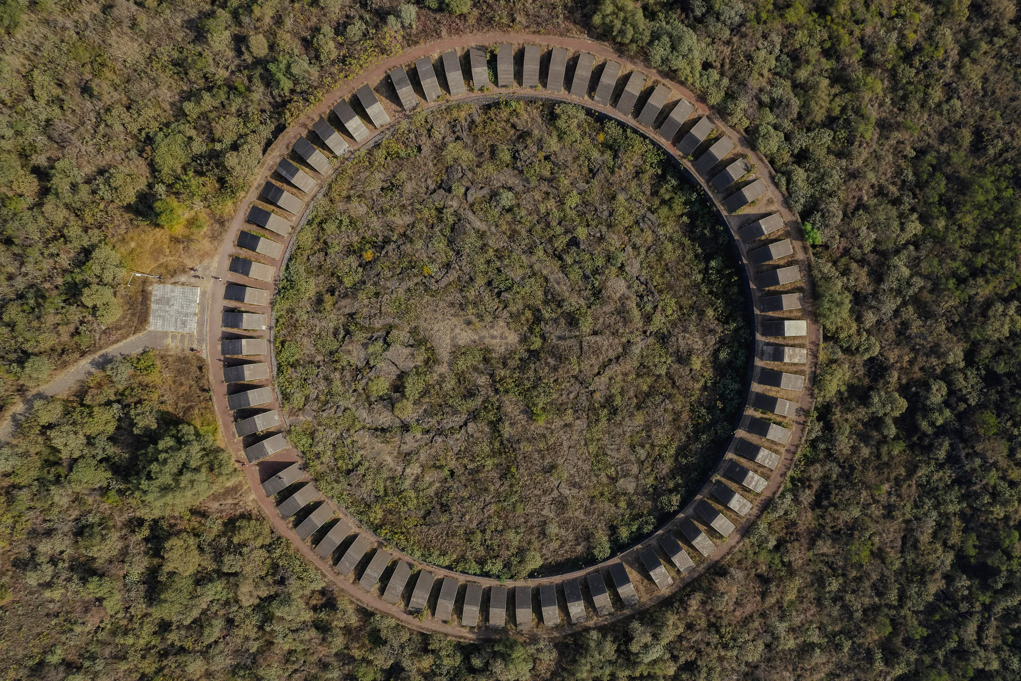 The Espacio Escultórico from directly above — a ring of sixty-four concrete wedges set into the black lava field at UNAM, scrub and forest all around it