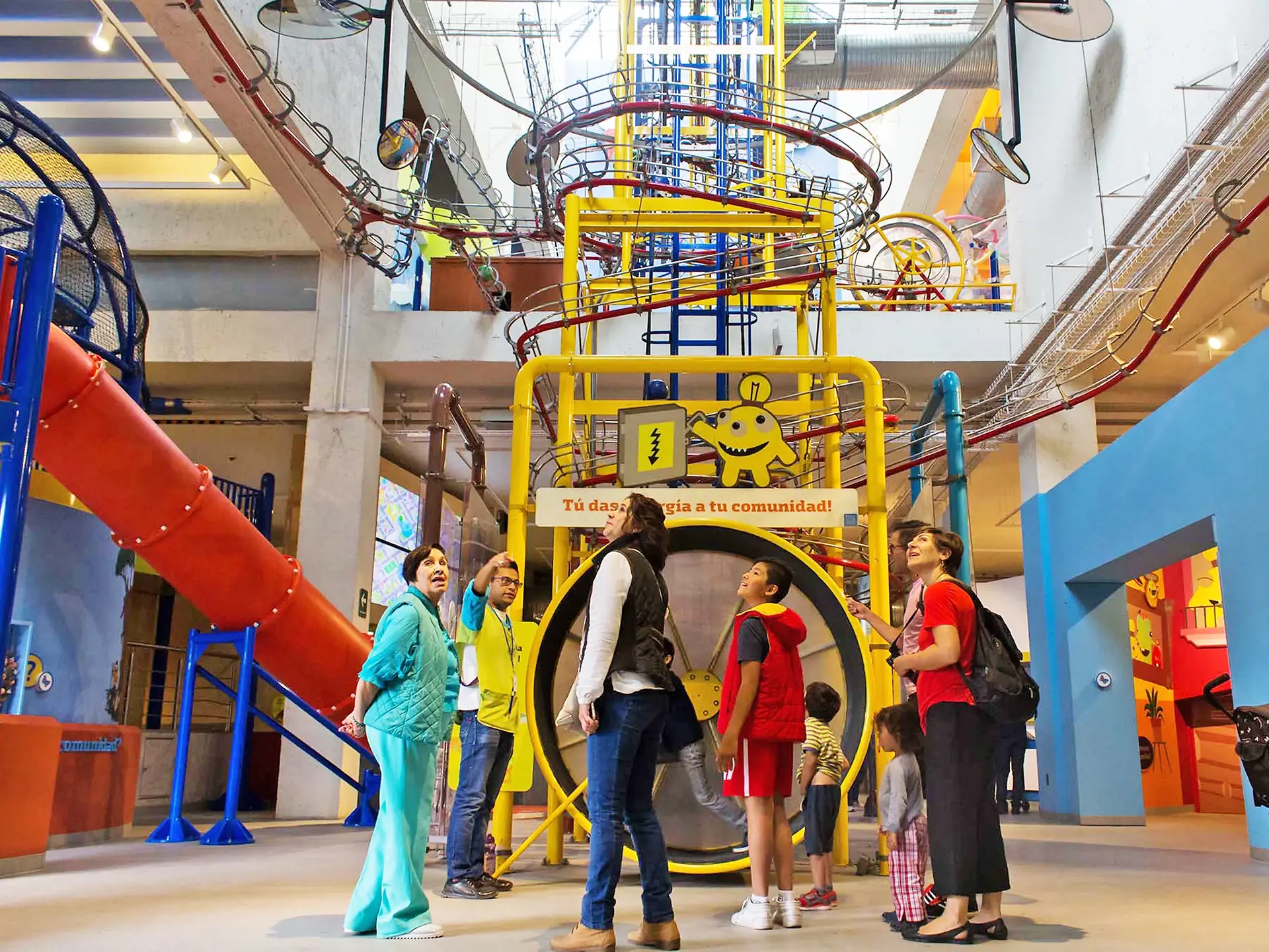 Museum atrium in primary colours — a yellow tower, a red tube slide and a ball run threading overhead, visitors gathered below