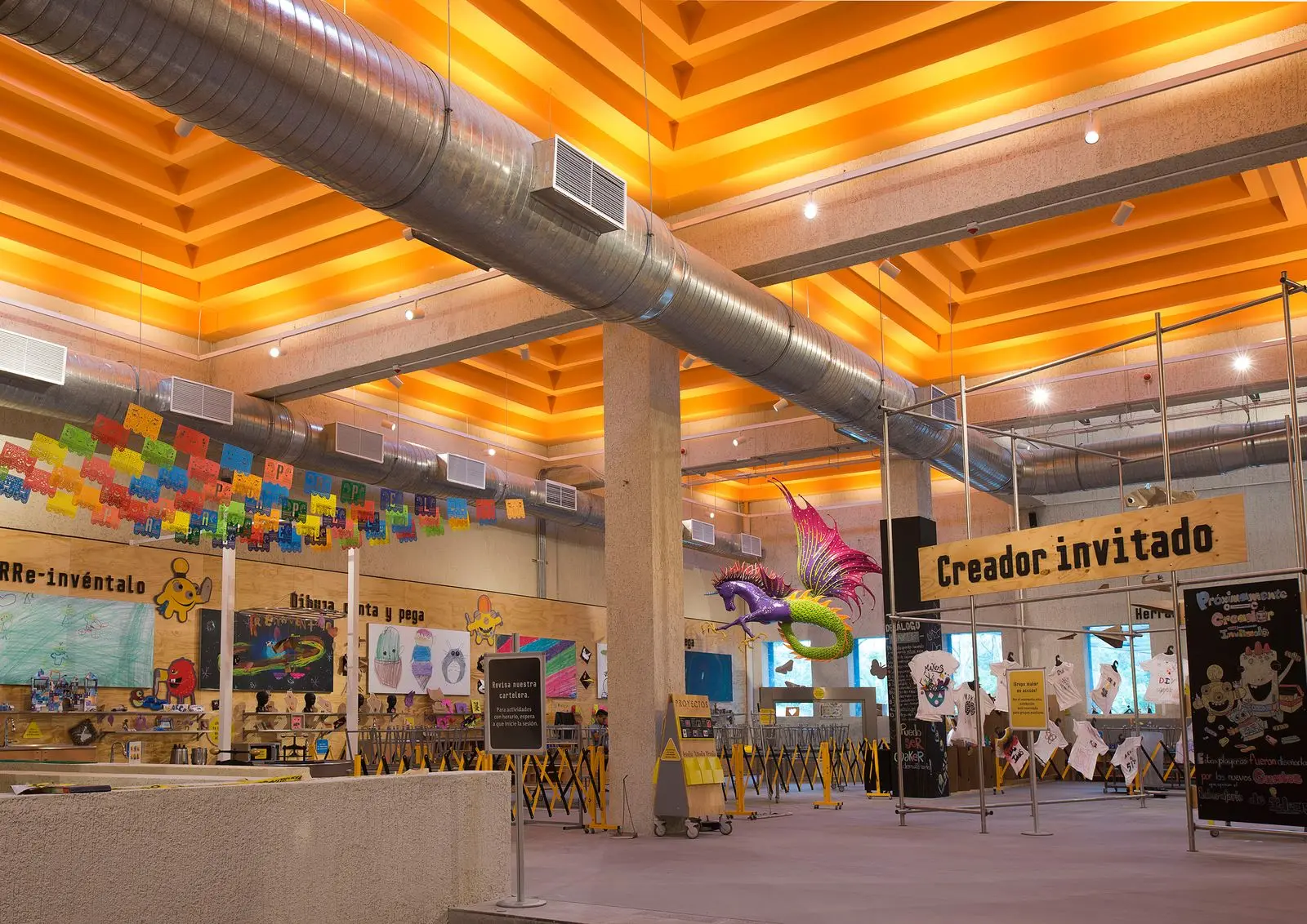 Workshop hall under an orange-lit slatted ceiling, a "Creador invitado" sign over benches and a hanging dragon sculpture
