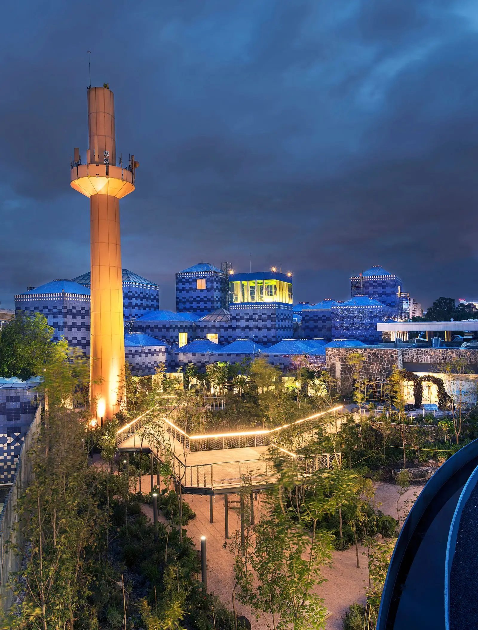 The museum at dusk — blue and white tiled domes under a storm sky, a tall orange chimney lit from below over a lantern-strung garden