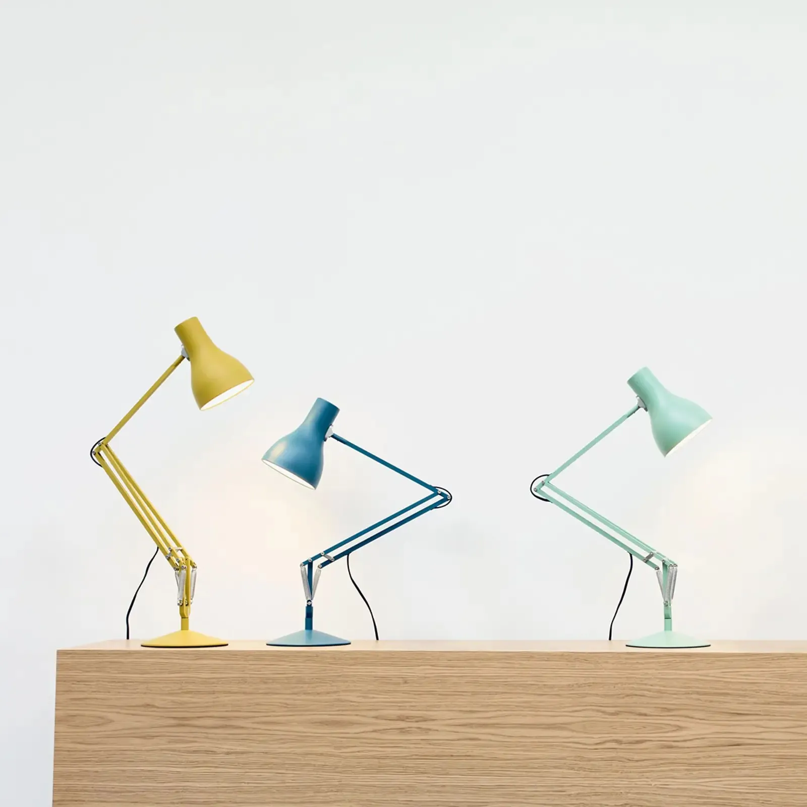 Three Anglepoise Type 75 desk lamps on an oak shelf — ochre, petrol blue and mint, arms folded at different angles