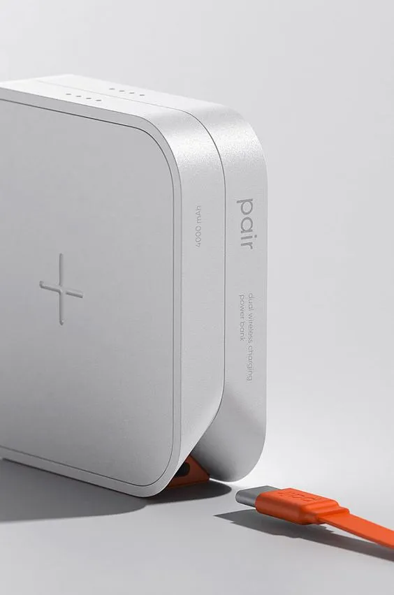 A silver "pair" wireless-charging power bank standing on a pale ground, an orange USB-C cable plugged into its foot