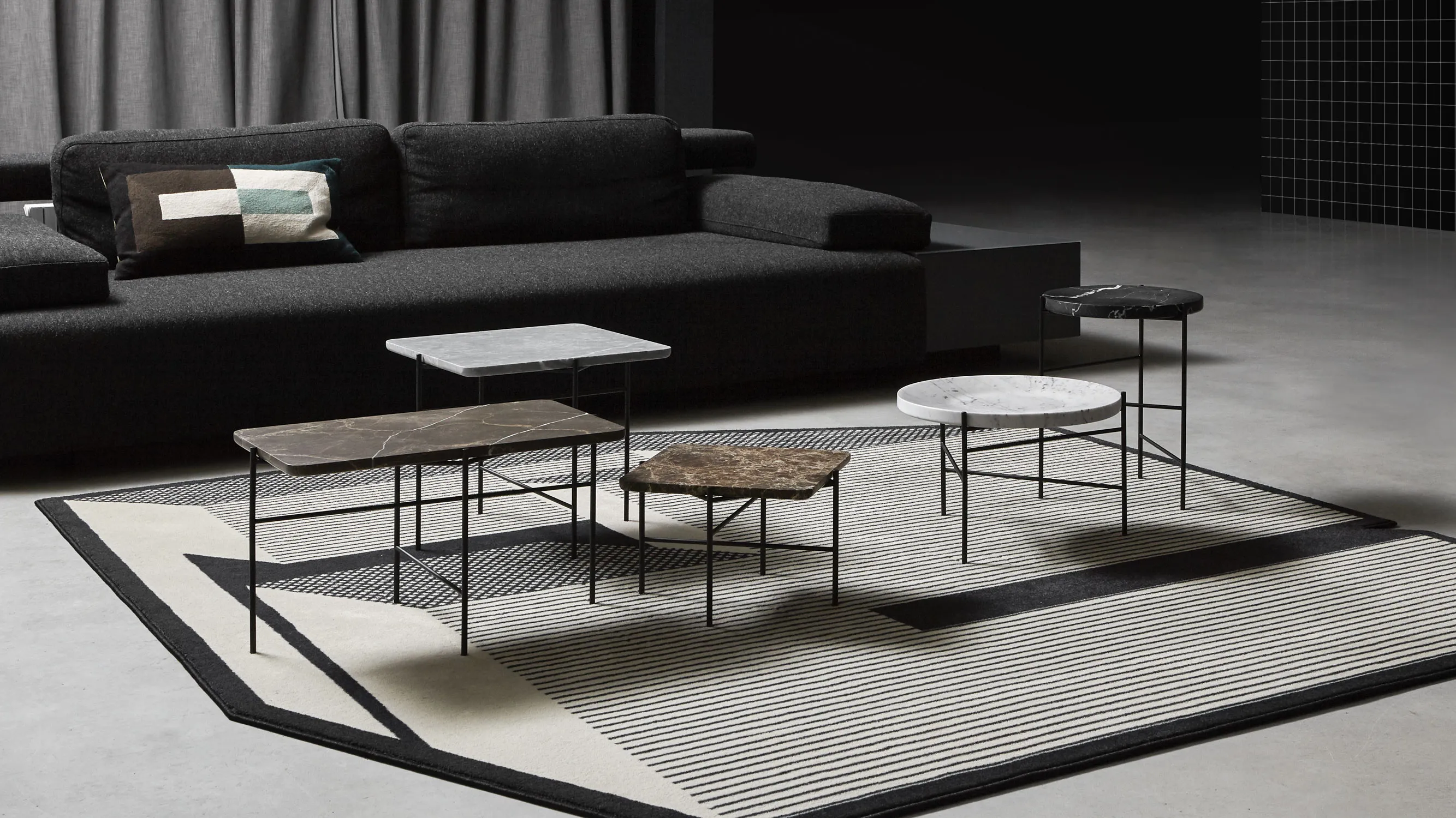 A low, dark living room — a black sofa behind a cluster of Paesaggi Sospesi tables at three heights, stone tops on thin black legs, on a pale striped rug
