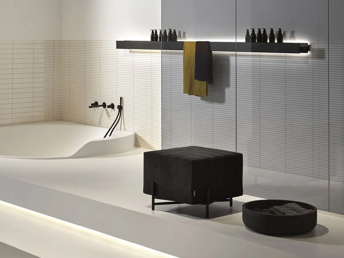A black pouf and a matching tray beside a sculpted white bath, in a pale bathroom with fluted wall panels