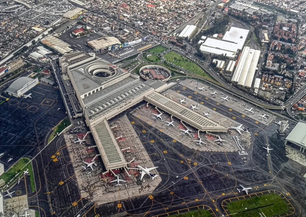 Aerial view of Mexico City airport — the terminal pier with aircraft parked along both sides, the city pressing up to the fence line